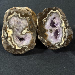 Op de afbeelding: Twee helften van een geode, met een levendig paars amethistkristal aan de binnenkant. De buitenste schil is een mix van bruin en beige, met een witte en crèmekleurige rand rond het kristal. De geode ligt op een donkere achtergrond.