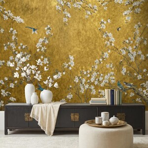 May include: A gold-toned wall mural featuring a floral design with white blossoms and small blue birds. A black cabinet with brass hardware holds white vases and books. A beige ottoman and rug complete the scene.