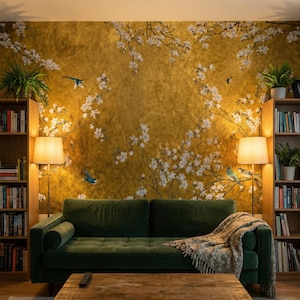 May include: A living room scene featuring a dark green velvet sofa, a wooden coffee table, and two wooden bookshelves. The focal point is a gold-toned wallpaper with white floral and bird designs. Two lamps provide warm lighting.