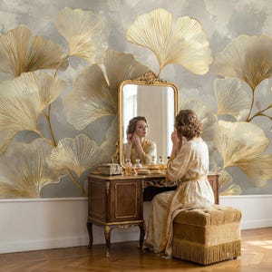 Puede incluir: Una mujer con una bata dorada está sentada en un tocador vintage, reflejada en un espejo con marco dorado. El tocador es de madera oscura con patas ornamentadas. El fondo presenta un mural de grandes hojas de ginkgo doradas.
