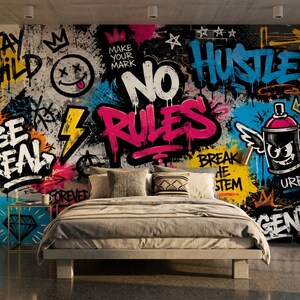 May include: A bedroom featuring a wall mural with vibrant graffiti art, including the text "STAY WILD," "BE REAL," "NO RULES," "HUSTLE," and "BREAK THE SYSTEM." A bed with neutral bedding sits in front of the wall, with a skateboard and shelves of sneakers.