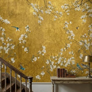 May include: A golden wallpaper with a floral design featuring white blossoms and small blue birds. The branches of the flowers extend across the wall. A wooden staircase and a table with books and a lamp are in the foreground.