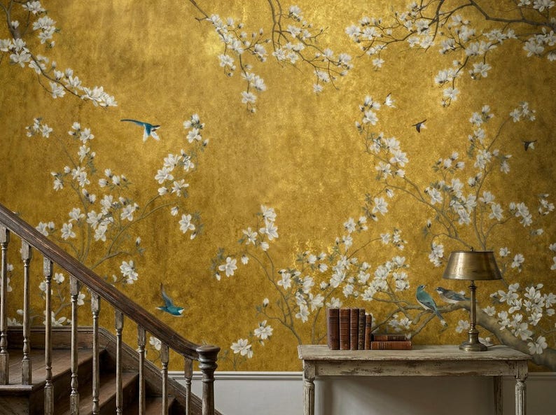 May include: A golden wallpaper with a floral design featuring white blossoms and small blue birds. The branches of the flowers extend across the wall. A wooden staircase and a table with books and a lamp are in the foreground.