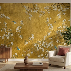 May include: A golden-hued wall mural featuring a floral design with white blossoms and branches. Blue birds are scattered throughout the scene. The mural is part of a room with a wooden cabinet, a beige armchair, and a wooden coffee table.