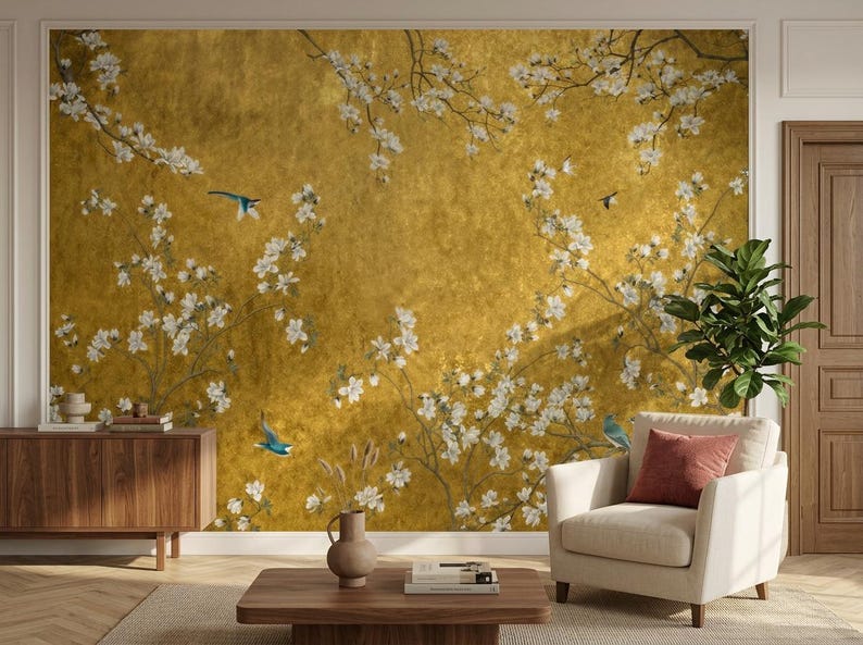 May include: A golden-hued wall mural featuring a floral design with white blossoms and branches. Blue birds are scattered throughout the scene. The mural is part of a room with a wooden cabinet, a beige armchair, and a wooden coffee table.