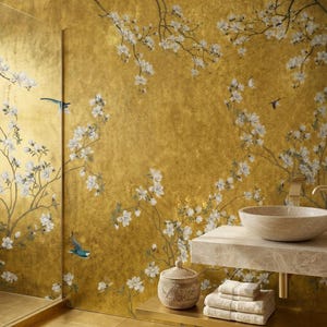 May include: A luxurious bathroom with a gold-toned wall featuring a floral and bird design. A marble countertop holds a stone sink and gold faucet. A woven basket, towels, and a glass shower door complete the elegant space.