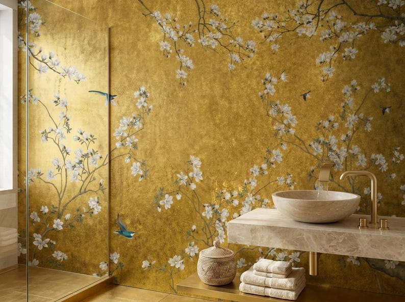 May include: A luxurious bathroom with a gold-toned wall featuring a floral and bird design. A marble countertop holds a stone sink and gold faucet. A woven basket, towels, and a glass shower door complete the elegant space.