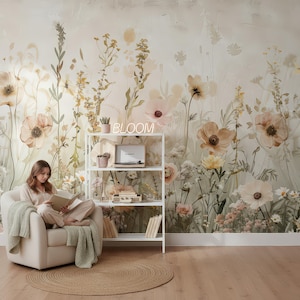May include: A woman sits in a cream-colored armchair, reading a book, with a green blanket draped over the arm. The background features a floral mural with large, soft-colored flowers. A white shelf holds a record player and decorative items, with a neon sign that says "BLOOM".