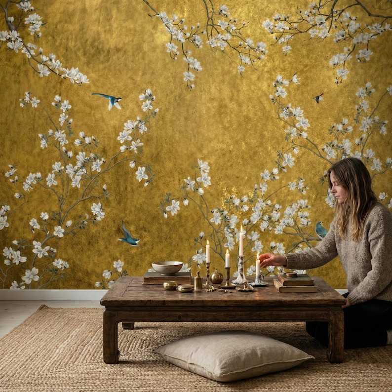 May include: A golden wallpaper with a floral design featuring white blossoms and blue birds. A wooden table with candles, books, and decorative items sits in front of the wallpaper. A beige rug and pillow are on the floor.