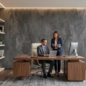 Op de afbeelding: Een modern directiekantoor met een groot houten bureau met een laptop, een computermonitor en een tablet. Een persoon in een grijs pak zit in een crèmekleurige lederen stoel, terwijl een andere persoon in een marineblauw pak ernaast staat.