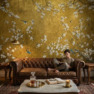May include: A living room scene featuring a brown leather Chesterfield sofa, a marble coffee table, and a gold-toned wall with a floral mural. A person is reading a book. A brass floor lamp and side tables complete the scene.