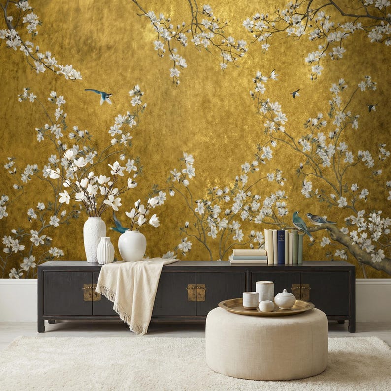 May include: A gold-toned wall mural featuring a floral design with white blossoms and small blue birds. A black cabinet with brass hardware holds white vases and books. A beige ottoman and rug complete the scene.