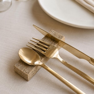 May include: A set of gold-toned stainless steel cutlery consisting of a spoon, a fork, and a knife resting horizontally on a rectangular, speckled beige stone block. The utensils feature a smooth, matte finish and are arranged side by side.
