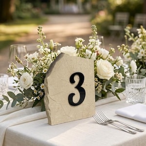 Beige stone wedding table number sign with black “3” displayed on an elegant outdoor reception table, surrounded by white floral arrangements, greenery, glassware, and soft natural sunlight in a garden setting.