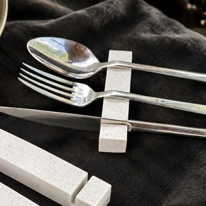 Könnte beinhalten: Ein Set aus vier rechteckigen, weiß gesprenkelten Besteckbänken aus Stein. Jede Besteckbank verfügt über eine lineare Einkerbung, um Utensilien sicher zu halten. Eine Besteckbank stützt einen Löffel, eine Gabel und ein Messer aus Edelstahl.