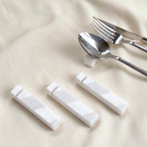 Könnte beinhalten: Ein Set aus weißen, rechteckigen Essstäbchenablagen aus Stein mit einer eingekerbten Oberfläche. Eine Ablage stützt einen Löffel, eine Gabel und ein Messer aus Edelstahl, während drei weitere Ablagen in einer Reihe darunter liegen.