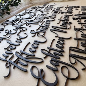 Peut inclure: Noms en bois noirs découpés au laser, en écriture cursive, disposés sur une surface claire. Les noms sont de différentes tailles et styles, parfaits pour la décoration personnalisée ou la signalisation d'événements. L'image présente une variété de noms.