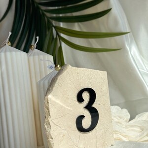 Beige stone wedding table number “3” with 3D  black acrylic lettering, styled with white ribbed candles, soft draped fabric, and a tropical leaf backdrop in a minimal, modern setting.