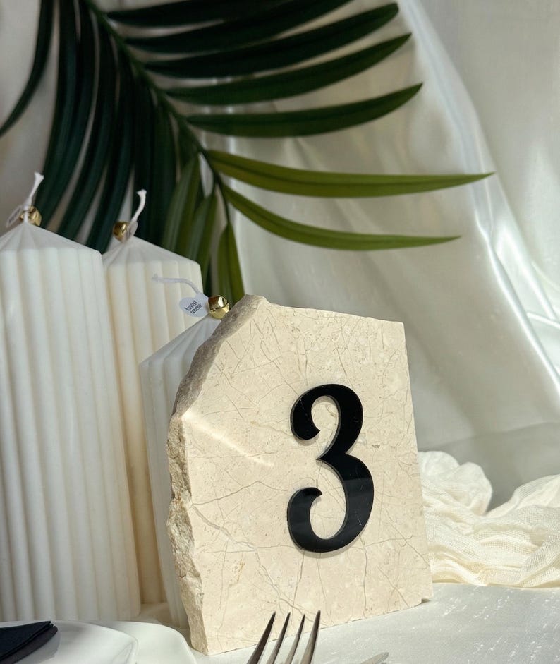 Beige stone wedding table number “3” with 3D  black acrylic lettering, styled with white ribbed candles, soft draped fabric, and a tropical leaf backdrop in a minimal, modern setting.