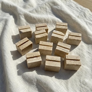 Può includere: Una collezione di dodici piccoli blocchi di pietra travertino beige di forma quadrata, ciascuno caratterizzato da una singola fessura orizzontale tagliata sulla superficie superiore, progettati per tenere carte o piccoli fogli in posizione verticale.