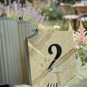 Travertine-style wedding table number “2” with 3d mirror silver acrylic lettering, styled beside white ribbed candles on a softly draped table, set against a romantic garden backdrop with pastel flowers and natural sunlight.
