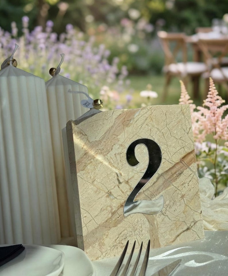 Travertine-style wedding table number “2” with 3d mirror silver acrylic lettering, styled beside white ribbed candles on a softly draped table, set against a romantic garden backdrop with pastel flowers and natural sunlight.