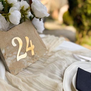 Travertine-style wedding table number “24” with gold mirror 3D acrylic lettering, styled with white florals on a soft draped fabric table, set in a sunlit outdoor reception with elegant tableware.