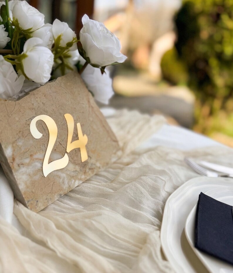 Travertine-style wedding table number “24” with gold mirror 3D acrylic lettering, styled with white florals on a soft draped fabric table, set in a sunlit outdoor reception with elegant tableware.