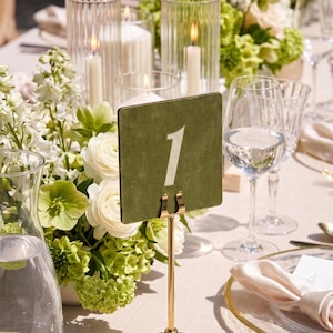 Könnte beinhalten: Eine elegante Tischdekoration mit einer grünen Tischnummerkarte mit der weißen Zahl 1, gehalten von einem goldenen Ständer. Der Tisch ist mit weißen Blumen, Kerzen und Glaswaren geschmückt.