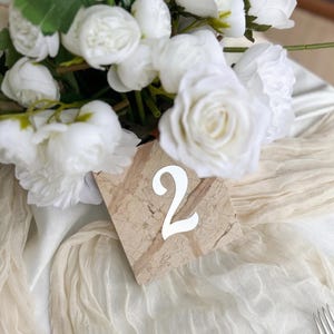 Marble-style wedding table number “2” with white acrylic lettering, placed beneath a soft white floral arrangement on a draped neutral fabric table setting with elegant textures and minimal styling.