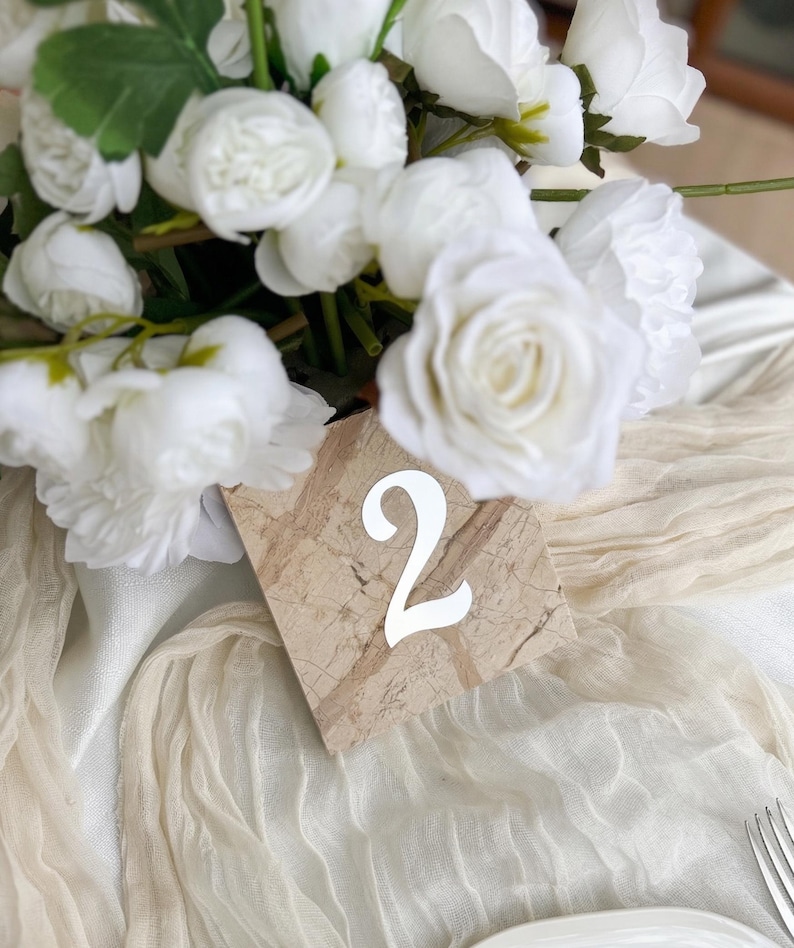 Marble-style wedding table number “2” with white acrylic lettering, placed beneath a soft white floral arrangement on a draped neutral fabric table setting with elegant textures and minimal styling.