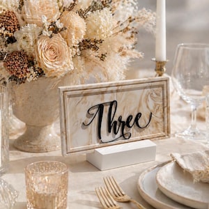 Op de afbeelding: Een gedekte tafel met een bloemstuk, kaarsen en een tafelnummer. Het tafelnummer is een bord met marmerpatroon met het woord "Three" in zwart script. Inclusief borden, glazen en goudkleurige vorken.
