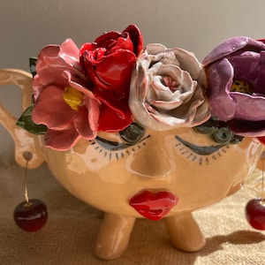 Könnte beinhalten: Ein beigefarbener Keramikübertopf in Form eines Gesichts mit geschlossenen Augen, langen Wimpern und leuchtend roten Lippen. Der Kopf ist mit modellierten roten, rosa, weißen und violetten Blumen verziert. Zwei kleine Kirschen hängen an goldenen Drähten.