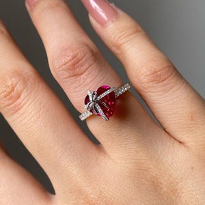 May include: A heart-shaped, ruby-red gemstone ring with a bow design. The ring features a silver band with small, clear accent stones. The gemstone is a vibrant red color, and the bow is made of smaller, clear stones.