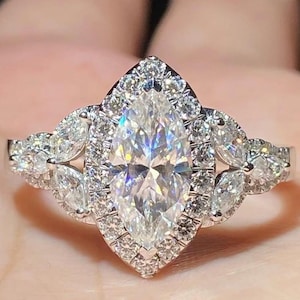 May include: A marquise-cut diamond engagement ring with a halo of smaller diamonds. The ring features a cluster of diamonds on each side of the main stone, set in a silver band. The ring is designed to sparkle and catch the light.