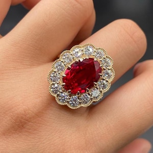May include: A gold ring featuring a large, oval-cut, deep red gemstone surrounded by a halo of smaller, clear gemstones. The ring's band has a detailed, textured design, and the setting is also gold.