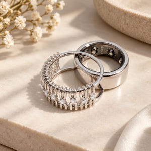 Puede incluir: Dos anillos de tono plateado, uno con una banda ancha y lisa con marcas internas y otro con un diseño tipo jaula engastado con pequeñas piedras claras. El anillo de jaula tiene barras verticales y una hilera de piedras.