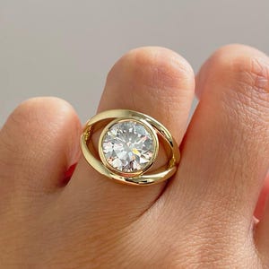 May include: A gold ring featuring a large, round, clear gemstone. The ring's band has a unique design, with a curved shape that encircles the gemstone. The ring is shown on a finger.