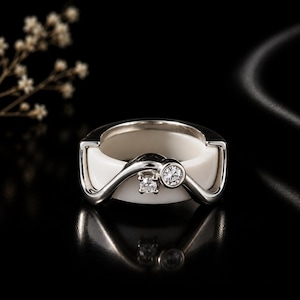 May include: A ring featuring a smooth white ceramic band encased in a polished silver-toned metal frame. The metal overlay forms a wave-like design across the front, accented by two round, clear, faceted stones set within the silver-toned structure.