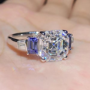 Puede incluir: Un anillo plateado con una gran piedra preciosa transparente de talla cuadrada como pieza central, flanqueada por dos piedras azules rectangulares más pequeñas. Pequeñas piedras transparentes adicionales están engastadas en los lados del anillo.