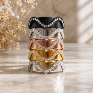 Puede incluir: Una pila vertical de cinco anillos, cada uno con un borde superior ondulado distintivo incrustado con pequeñas piedras redondas transparentes. Los anillos tienen acabados en negro mate, plata pulida, oro rosa, oro amarillo y tonos de plata pulida.