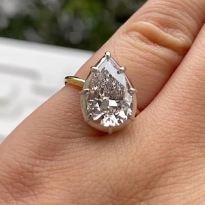 May include: A diamond ring featuring a pear-cut stone, set in a gold band. The diamond is held in a platinum or white gold setting. The ring is displayed on a finger, with a blurred green backdrop.