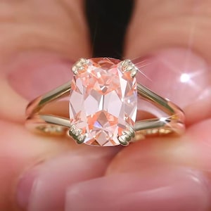 May include: A gold ring featuring a large, cushion-cut, peach-colored gemstone held by four prongs. The band has a split-shank design that tapers toward the center stone, creating a polished, metallic finish that reflects light across the surface.