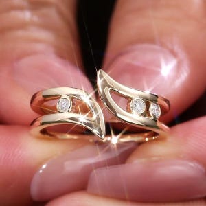 May include: A gold bypass ring featuring a split shank design with three small, round, clear stones set in bezel settings. The metal has a polished, reflective finish that catches the light, creating a bright, sparkling effect on the surface.