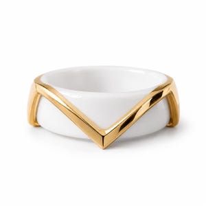 Peut inclure: Une bague composée d'un anneau en céramique blanche lisse rehaussé d'une superposition en métal doré poli. L'élément métallique forme un motif en chevron en V distinct qui enveloppe l'avant de l'anneau blanc, créant un contraste entre les matériaux.