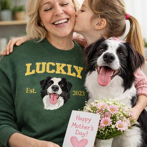 以下が含まれることがあります： 金色の文字で「LUCKLY」と犬のグラフィックが描かれた濃い緑色のスウェットシャツ。スウェットシャツには「Est. 2023」の文字も入っています。犬、花、そして「Happy Mother's Day!」のカードも画像にあります。