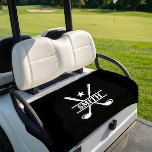 May include: A black golf cart seat cover with a white graphic design featuring crossed golf clubs, a star, and the name "SMITH". The golf cart has white seats and a white body, set against a green golf course.