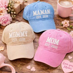 以下が含まれることがあります： ベージュ、水色、ピンクの野球帽が3つ。各帽子には白い刺繍で「MOMMY EST. 2018」、「MAMAN EST. 2018」、および「MOM EST. 2018」と書かれています。帽子は、花とピンクのマグカップが置かれた木製の表面にあります。
