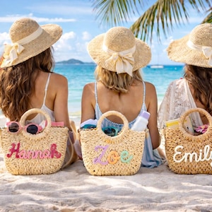 Puede incluir: Tres mujeres sentadas en una playa, cada una con un bolso de mano de paja. Los bolsos están personalizados con los nombres "Hannah", "Zoe" y "Emily" en letras coloridas. Cada mujer lleva un sombrero de paja con una cinta.
