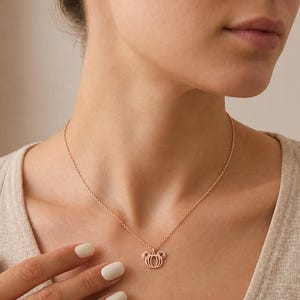 May include: A rose gold-toned necklace featuring a delicate chain and a small, open-work pendant shaped like a stylised pumpkin with rounded, ear-like protrusions at the top, resting against a neutral-coloured ribbed knit top.